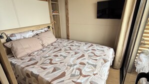 Un lit avec une couvre-lit à motifs, une tête de lit en bois, un luminaire suspendu et un téléviseur fixé au mur.