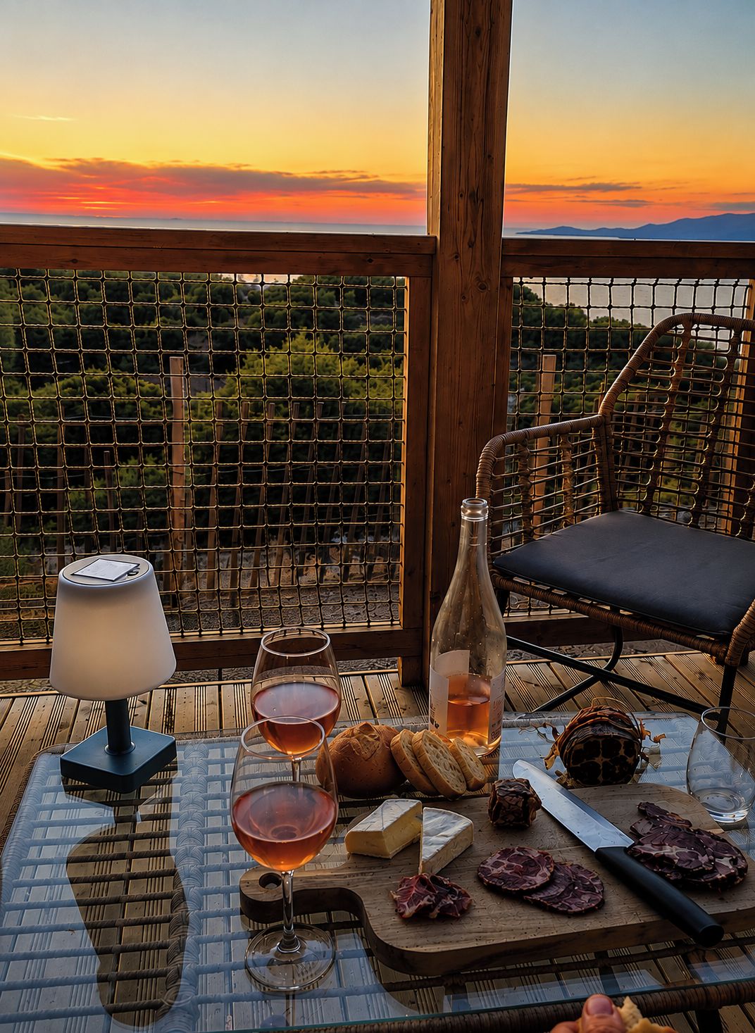 Een tafel gedekt met wijnglazen, een fles, kaas en charcuterie op een balkon met uitzicht op de zonsondergang en een wijngaard.