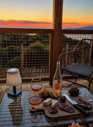 Une table dressée avec des verres à vin, une bouteille, du fromage et de la charcuterie, sur un balcon offrant une vue sur le coucher de soleil et les vignes.