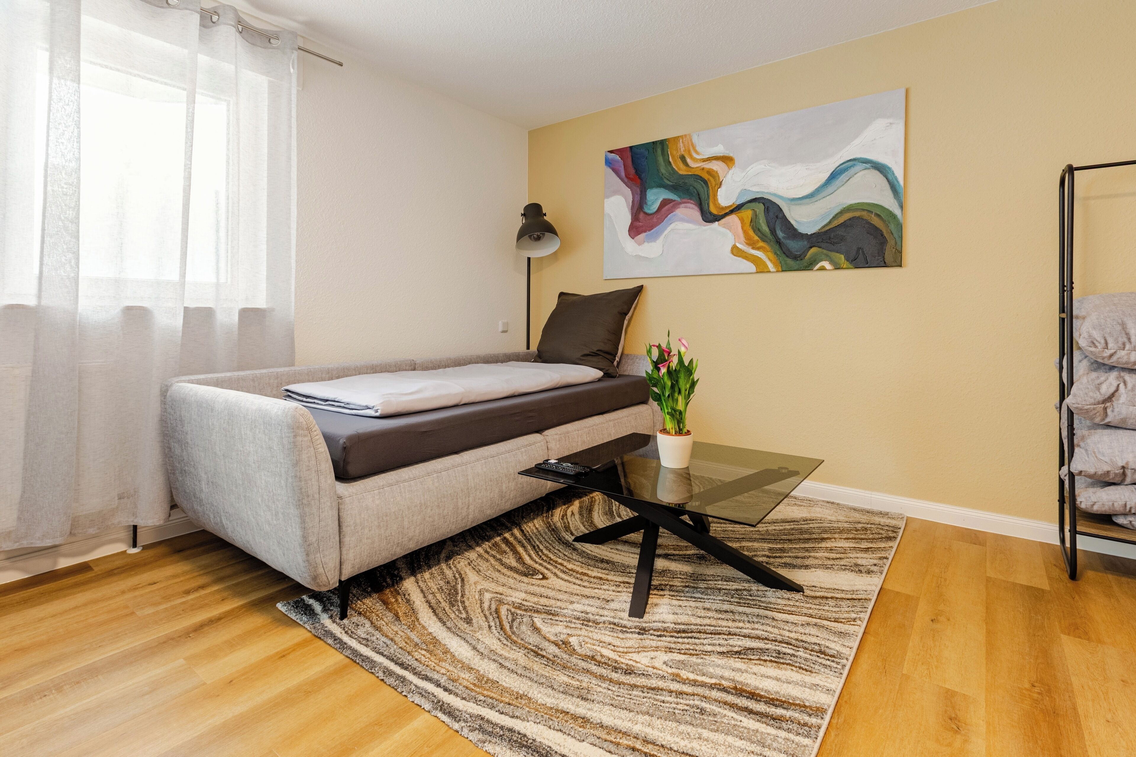 A modern bedroom with a sofa bed, a glass coffee table, a painting on the wall, and a potted plant.
