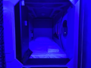 Une pièce étroite et faiblement éclairée, avec un lit simple, une petite table et un luminaire fixé au mur.