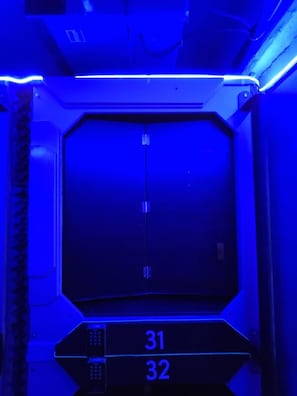 A room with a large, dark, rectangular window, two control panels with numbers 31 and 32, and a blue ambient light. - Hotel Capsula Condesa (Mexico City)