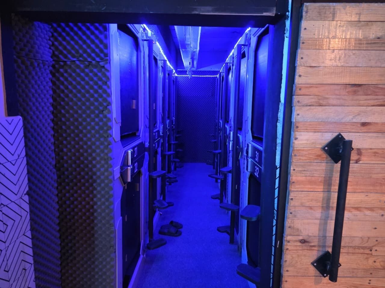 Un pasillo estrecho con hileras de equipos, posiblemente para producción de audio o video, con una puerta de madera a la derecha.