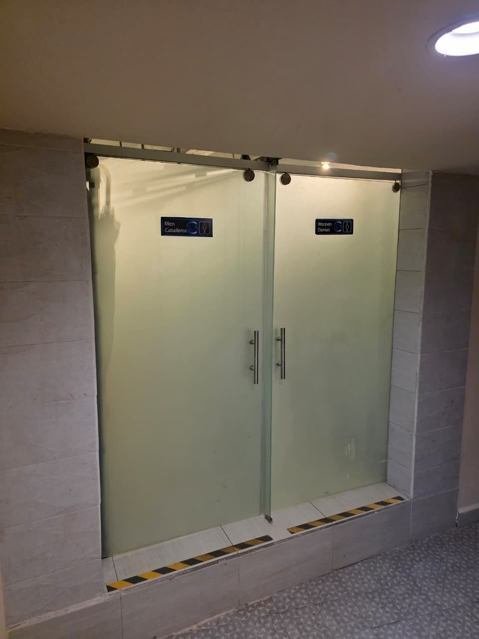 Eine Doppeltür mit frostigem Glasfenster, ein Schild mit den Hinweisen 'Herren' und 'Damen' Toiletten sowie ein gefliester Boden mit gelb-schwarzem Warnstreifen.