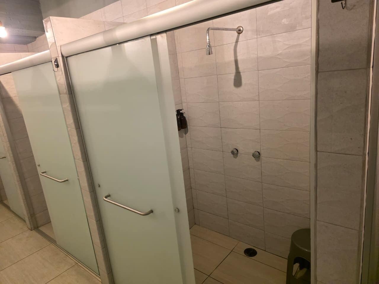 Una hilera de cabinas de baño con una zona de duchas; cada cabina tiene una puerta con manija, y el suelo está cubierto de azulejos.