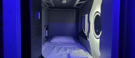 Una habitación pequeña con una cama individual, un ventanal y una lámpara circular.
