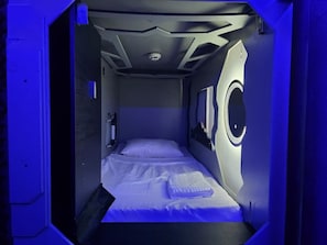 A compact room with a single bed, a small window, and a circular light fixture. - Hotel Capsula Condesa (Mexico City)
