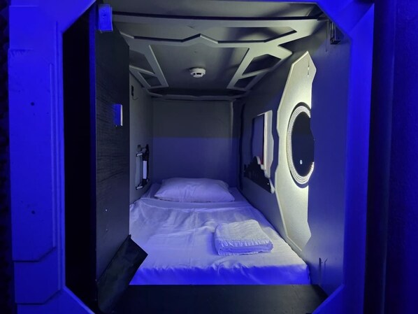 A compact room with a single bed, a small window, and a circular light fixture.