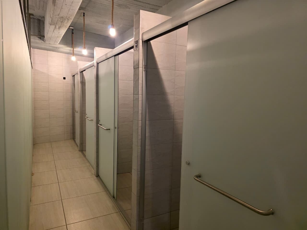 Een gang met deuren naar individuele kamers, een betegelde vloer en een plafond met blootliggende balken en hangende lampen.
