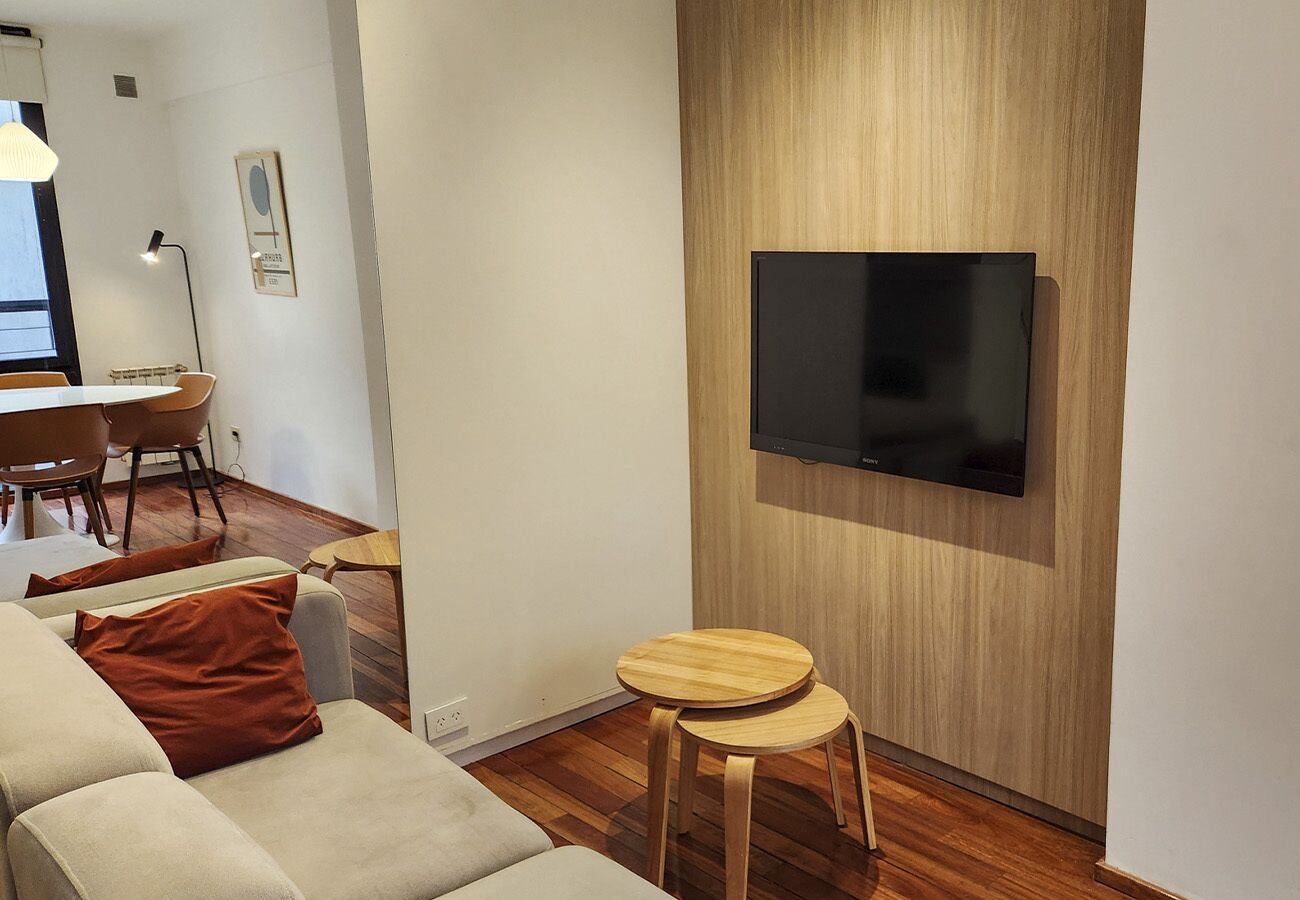 A modern living room with a flat-screen TV mounted on a wooden wall, a sofa with a brown pillow, and a small wooden stool.