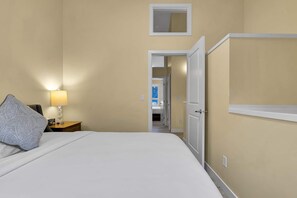 Una camera d'albergo con un letto, un comodino con una lampada, una porta che dà su un corridoio e un piccolo specchio a parete.