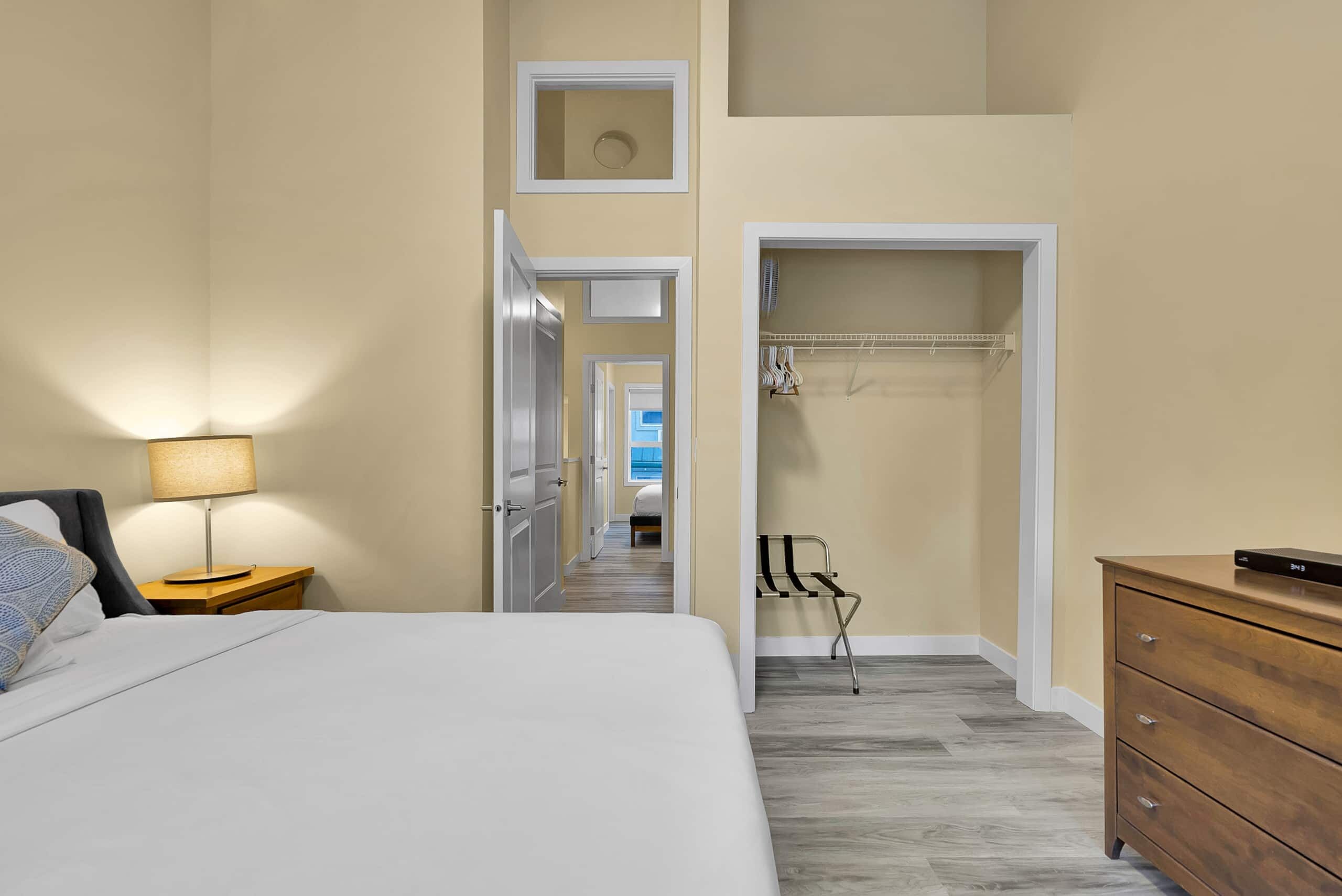 Habitación de hotel con cama, mesita de noche, lámpara, cómoda de madera y vestidor con ropa colgada.