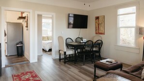 Una sala de estar con alfombra roja, un sofá marrón, una mesa de centro, un televisor montado en la pared y un comedor con sillas y una mesa.