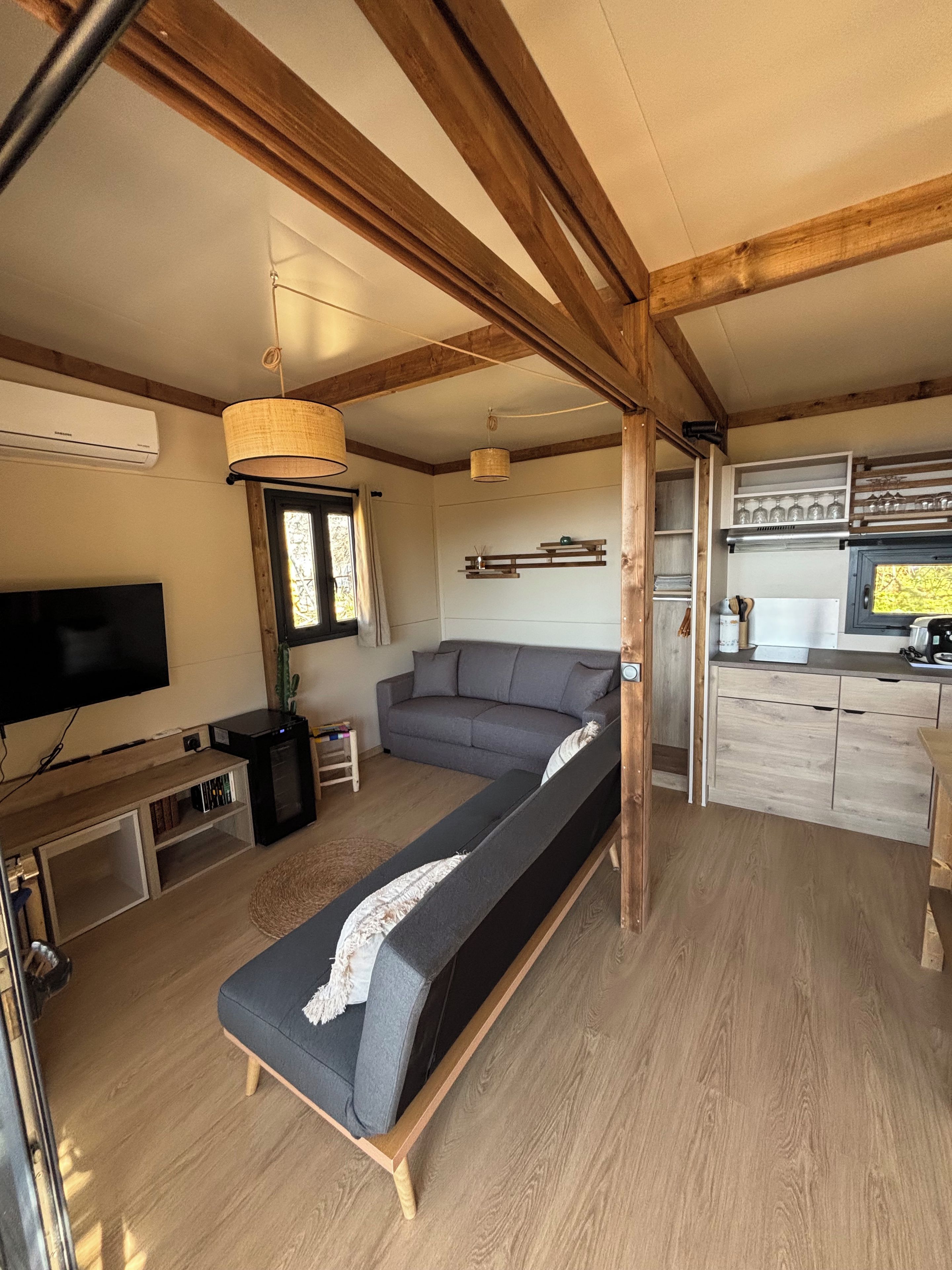 Interior moderno de una casa rodante con sofá, televisor, kitchenette y vigas de madera.