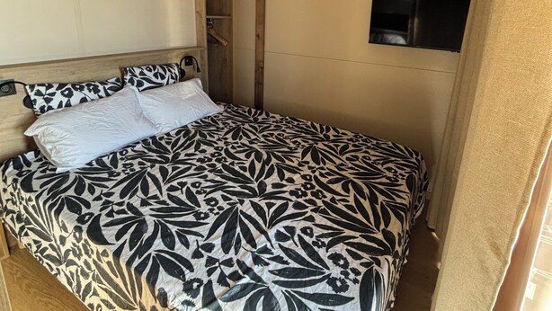 Ein Bett mit gemustertem Überwurf, ein an der Wand befestigter Fernseher und Holzbalken an der Decke.