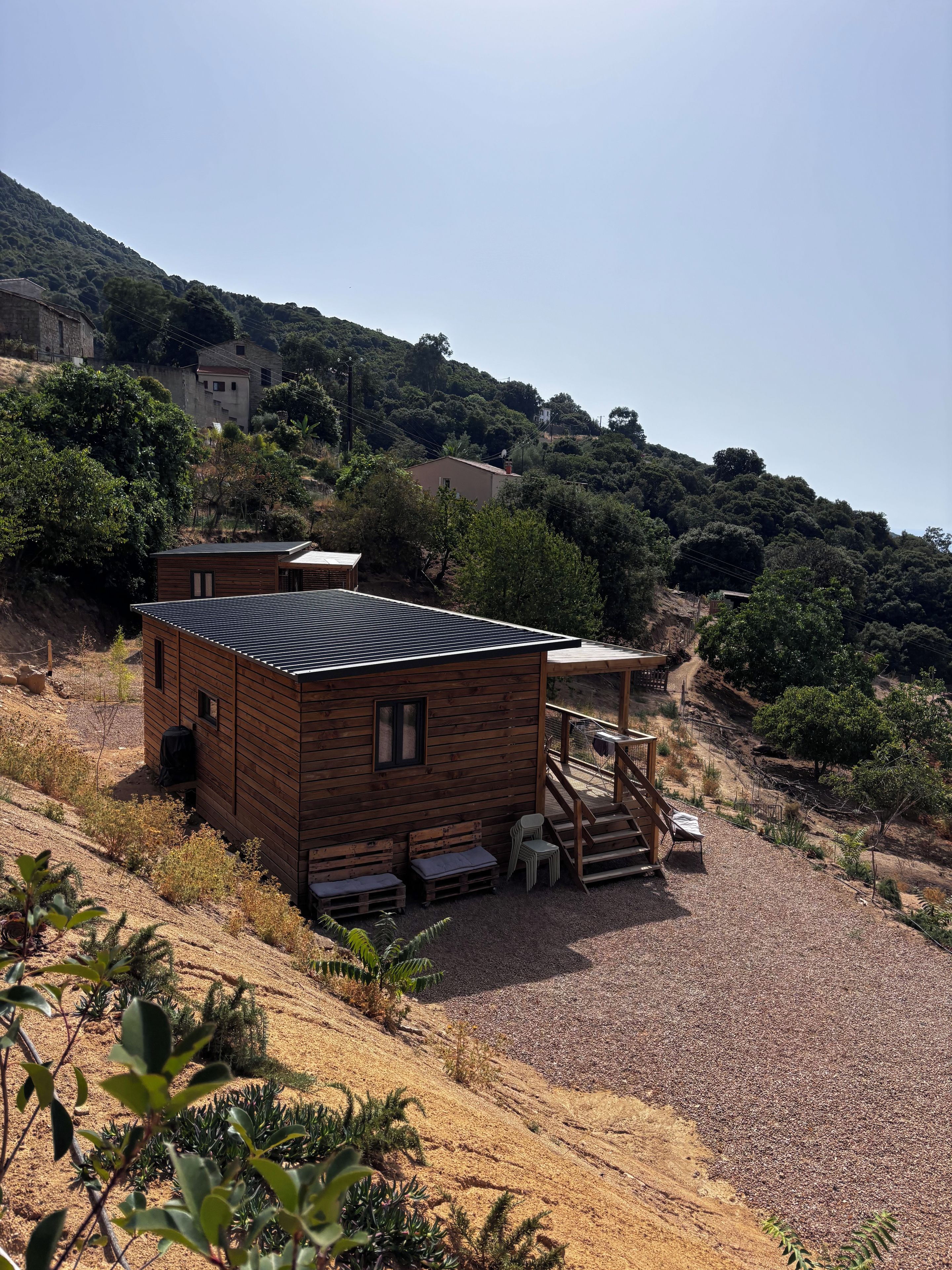 A wooden cabin with a deck, surrounded by a gravel area and vegetation.