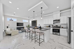 A modern kitchen with white cabinets, a central island with bar stools, a large TV, and a dining area with a sofa and chairs. - Executive Home, Office, Galleria & NRG (Houston)