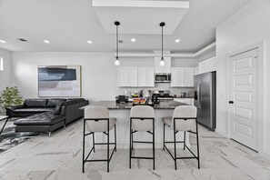 A modern kitchen with white cabinets, a stainless steel refrigerator, and a bar area with three stools. - Executive Home, Office, Galleria & NRG (Houston)