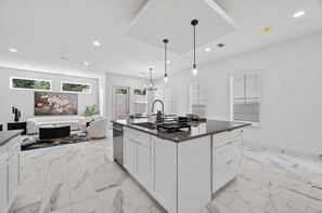 Cozinha moderna com armários brancos, uma ilha central, iluminação pendente e uma área de jantar com sofá e mesa de centro.