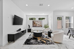 A modern living room with a flat-screen TV, white sofas, a coffee table, and a large painting. - Upscale Home, Office, Galleria & NRG (Houston)