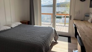Ett sovrum med en säng, ett nattduksbord, ett skrivbord i trä, en stol, ett fönster med utsikt över en sjö och en dörr som leder ut till en uteplats.