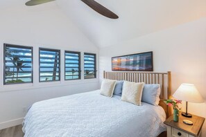 A bedroom with a bed, a nightstand, a lamp, a painting, and three windows with a view of the beach. - ALL NEW Oceanfront 2 storey comfortably sleeps 6-7 (Lihue)
