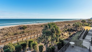 Vista de la playa con un muelle de madera, palmeras y un patio de hormigón con mesa y sillas.