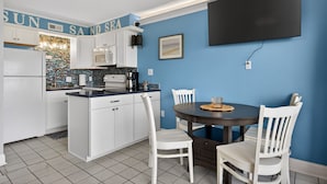 Una cocina con armarios blancos, una pared azul, un televisor de pantalla plana sobre la mesa de comedor y piso de baldosas.