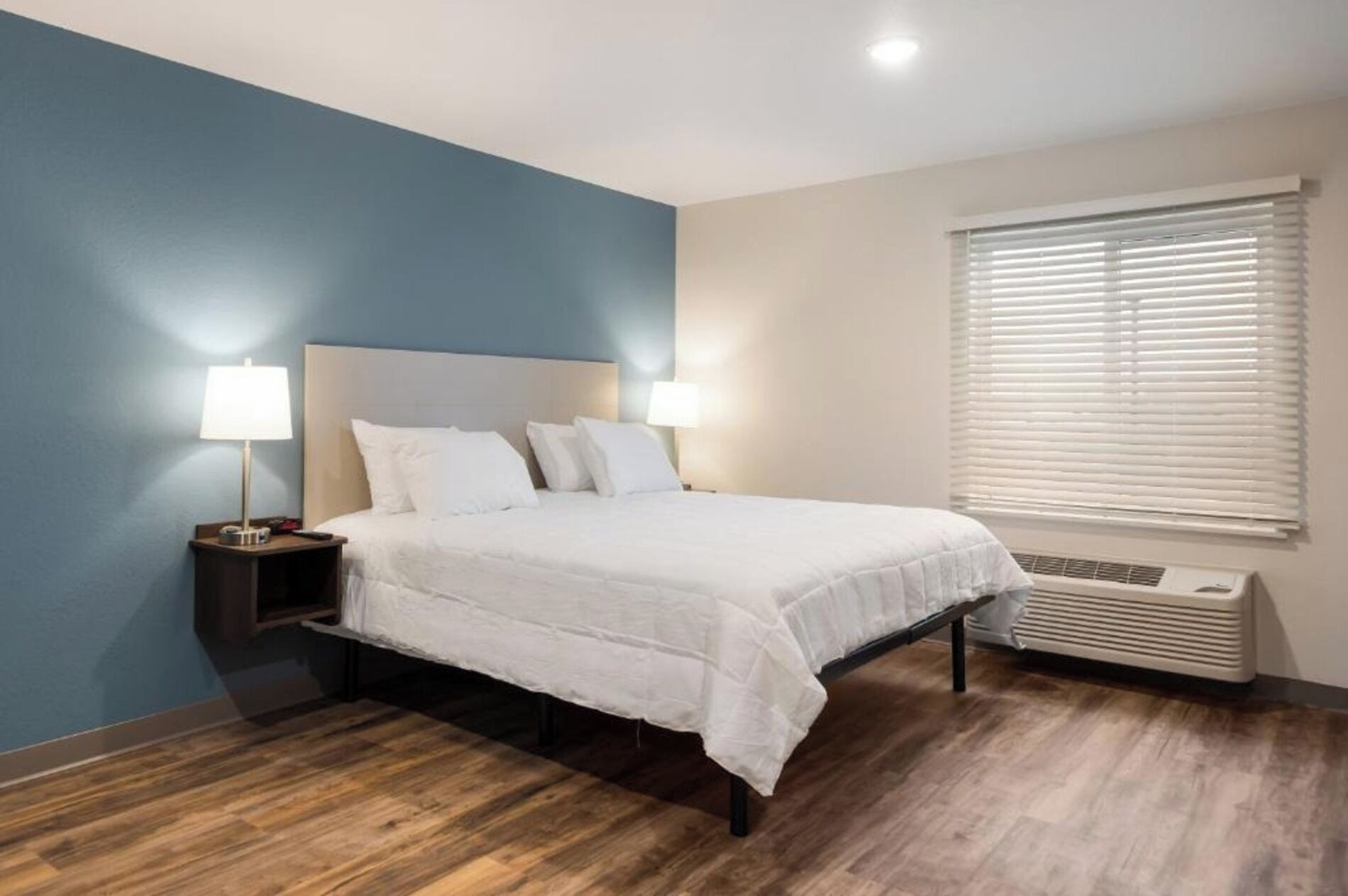A bedroom with a bed, two bedside lamps, a window with blinds, and a wall-mounted air conditioner.
