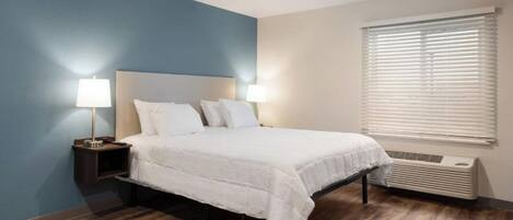 A bedroom with a bed, two bedside lamps, a window with blinds, and a wall-mounted air conditioner.