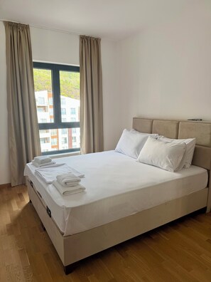 Een net opgemaakt bed met witte lakens en kussens, een houten vloer en een raam met gordijnen.