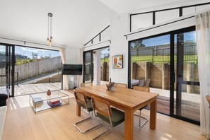Ein moderner Essbereich mit einem Holztisch und -stühlen, einem Glastisch und Blick auf eine Holzterrasse und einen Garten durch große Schiebeglastüren.
