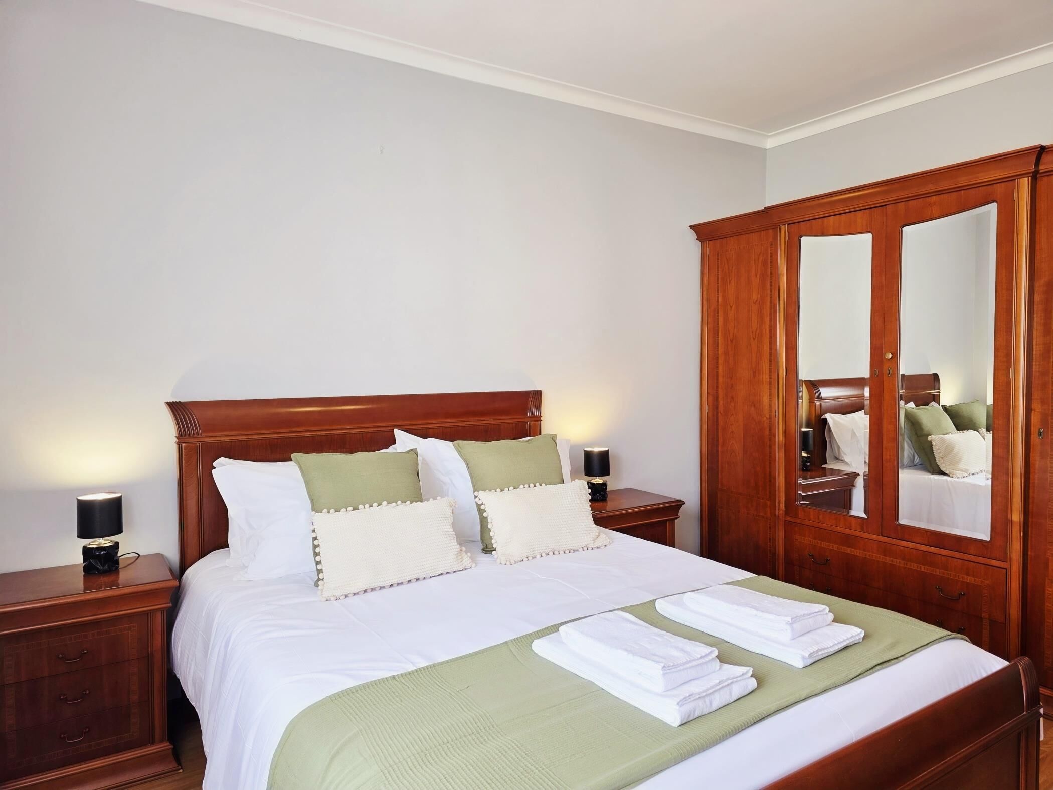 A neatly made bed with white linens and a green throw, wooden nightstands with lamps, and a large mirror cabinet.