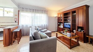 A living room with a sofa, a coffee table, a TV stand, and a kitchen area with a counter and stools. - Portas de Melgaço Apartment | Minho | The northernmost town in Portugal (Melgaço)