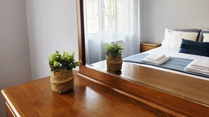 A neatly made bed with pillows, a wooden nightstand, a window with sheer curtains, and two small potted plants on a wooden dresser. - Portas de Melgaço Apartment | Minho | The northernmost town in Portugal (Melgaço)