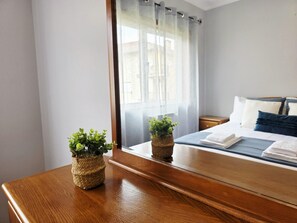 Ein ordentlich bezogenes Bett mit Kissen, ein Nachttisch aus Holz, ein Fenster mit transparenten Vorhängen und zwei kleine Topfpflanzen auf einer Holzkommode.