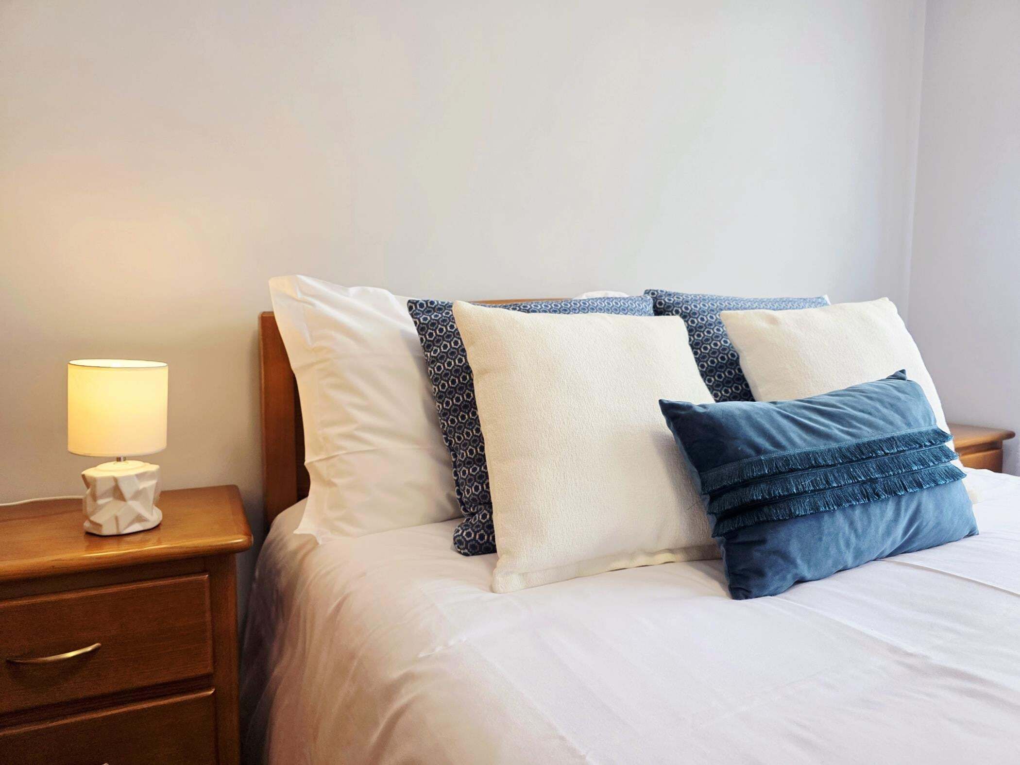 A neatly made bed with white and patterned pillows, a bedside lamp, and a wooden nightstand with drawers.
