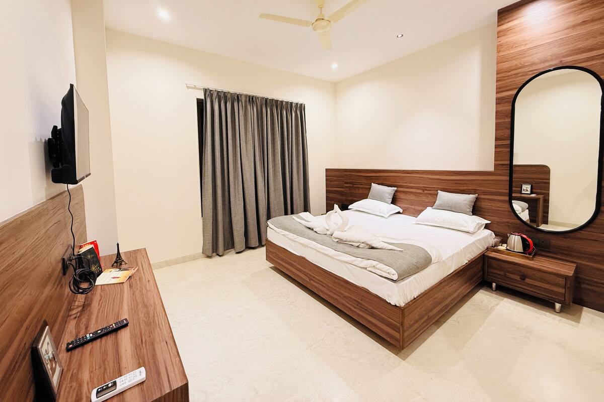 A modern hotel room with a wooden bed, a flat-screen TV, a ceiling fan, a large mirror, and a bedside table with a lamp.