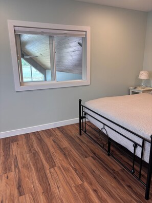 A bedroom with a bed, a nightstand, a lamp, and a window with a view of the outdoors. - Brand New 2 Bedroom Lake Home in Charming Cumberland, WI (Cumberland)
