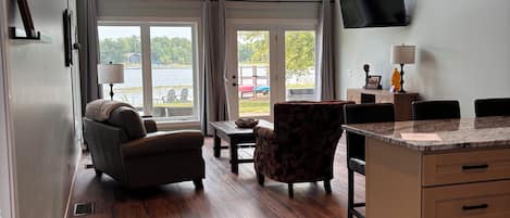 A spacious room with a high ceiling, large windows, a flat-screen TV, a sofa, a chair, and a kitchen area with a countertop and drawers.
