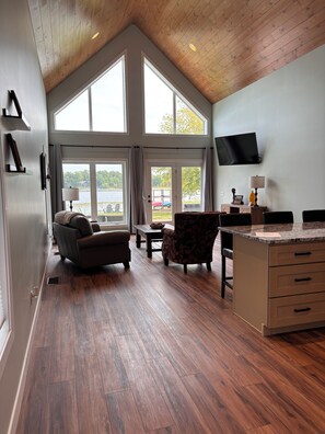 A spacious room with a high ceiling, large windows, a flat-screen TV, a sofa, a chair, and a kitchen area with a countertop and drawers. - Brand New 2 Bedroom Lake Home in Charming Cumberland, WI (Cumberland)
