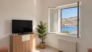 A living room with a TV on a wooden cabinet, a potted plant, a window with a view of a coastal town, and a radiator. - 3 bedroom beach front apartment in Stomorska (Stomorska)