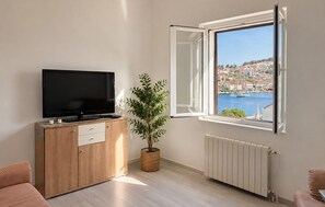 Ett vardagsrum med en tv på ett träskåp, en krukväxt, ett fönster med utsikt över en kuststad och ett element.