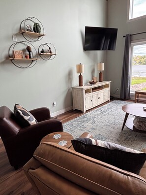 A living room with a brown leather sofa, two lamps, a television, and a view of a lake through a window. - Brand New 3-Bedroom Lakefront Home with PICKLEBALL and PONTOON rental option (Cumberland)
