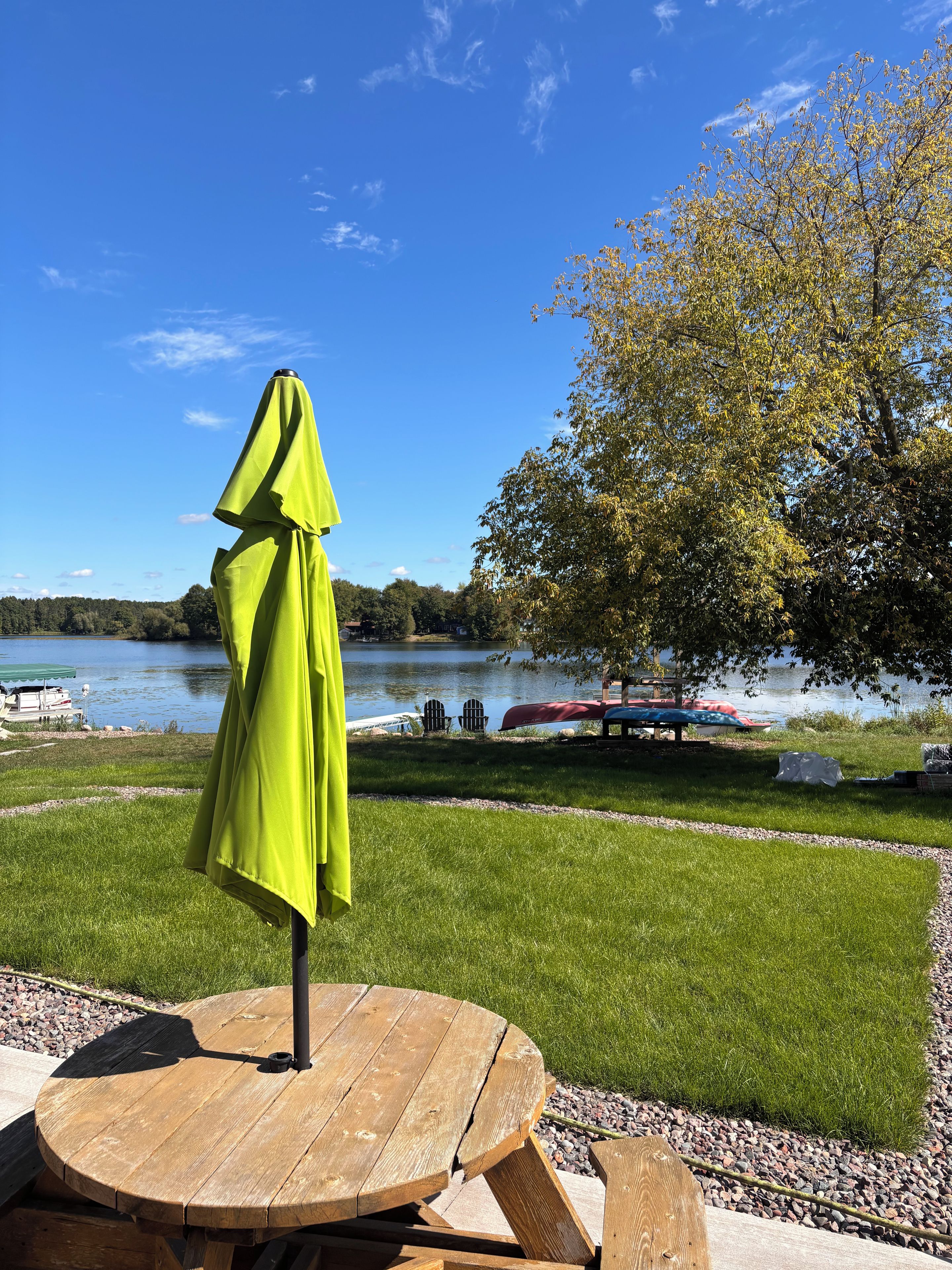 A wooden table with a green umbrella, a grassy area, and a lake with boats in the background.