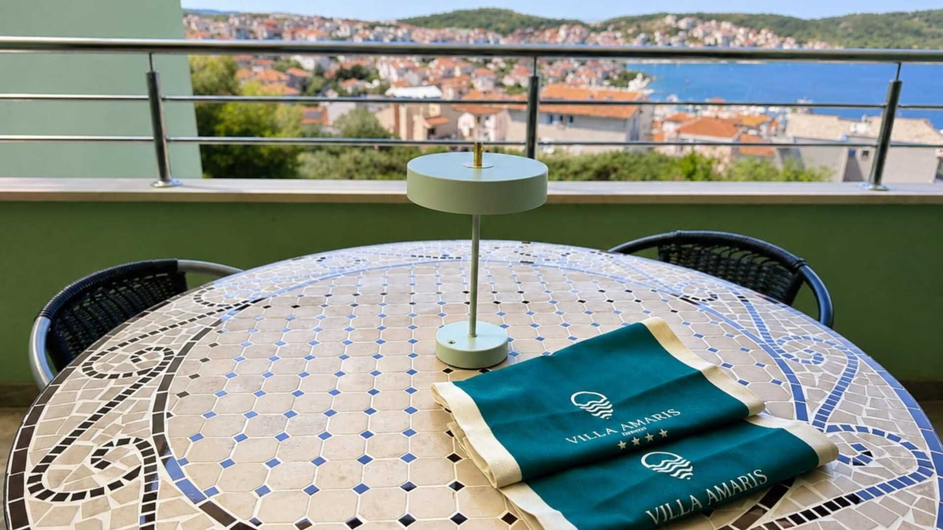 Et rundt bord med et mosaikdug, to stole, en bordlampe og et grønt klæde med 'Villa Amaris' logo.