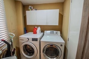 A laundry area with two washing machines, a cabinet above, and a window with blinds. - Sonoran Spa W102 Ground Floor 2BR/2ba (Puerto Peñasco)