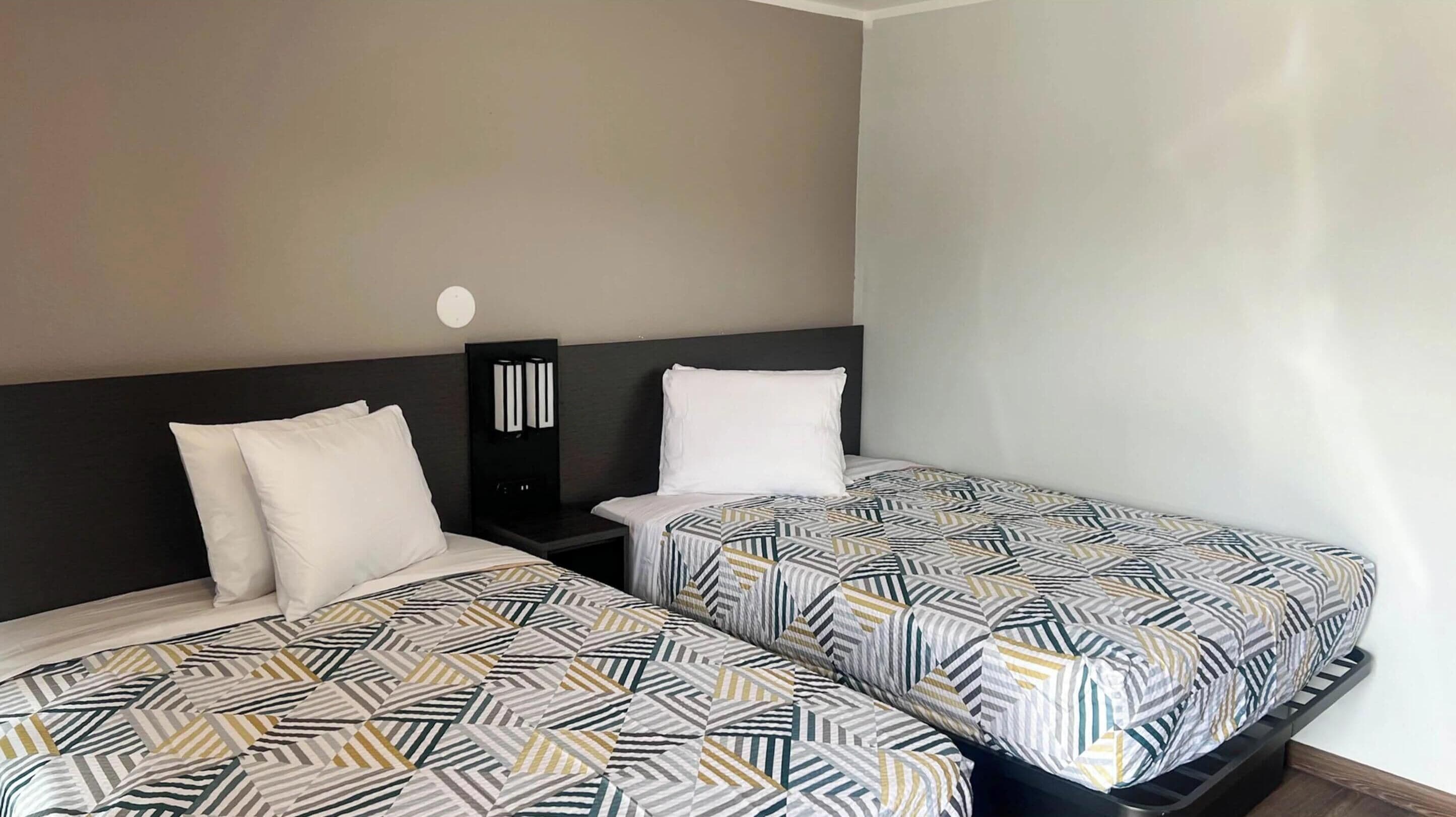 A hotel room with two beds, each with patterned bedding, a nightstand, and a wall-mounted light fixture.