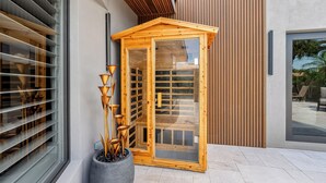 Een houten sauna met glazen deuren, een bloempot met hoge, slanke planten, en een modern gebouw met houten lamellen en een groot raam.