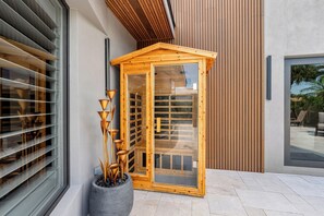 Eine Holzsauna mit Glastüren, ein Blumentopf mit hochwachsenden Pflanzen und ein modernes Gebäude mit Holzlamellen und einem großen Fenster.